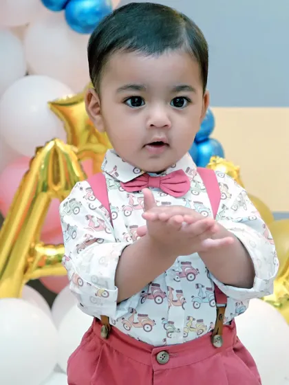 A well-dressed little boy claps his hands with excitement in front of a balloon-filled birthday backdrop. This is a perfect example of a classic birthday portrait.
