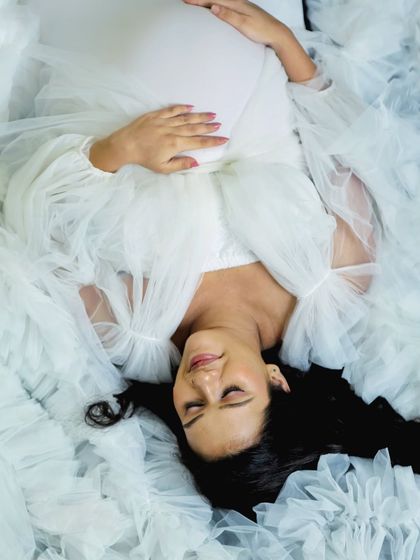 A serene, artistic shot from above. The mom-to-be lies peacefully, enveloped in the soft, cloud-like fabric of a white ruffled gown, creating a truly angelic portrait.