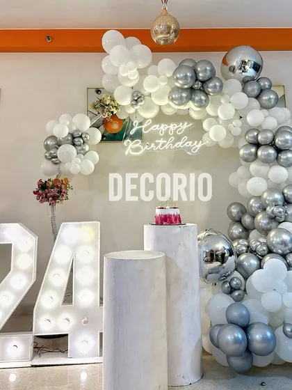 A chic 24th birthday setup with a silver and white color scheme. The decor includes a balloon arch, '24' marquee numbers, and pedestals for the cake, creating a clean and modern look.