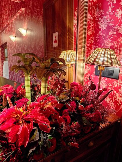 This entryway arrangement sets a dramatic tone with a profusion of red flowers against a bold, patterned wallpaper. The wicker lamp and miniature palm tree models add a touch of tropical, eclectic charm.