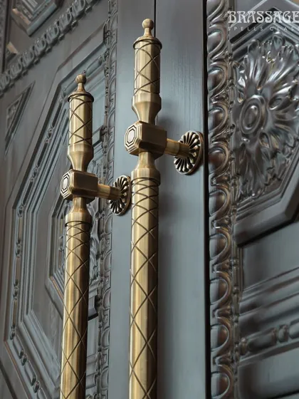 Our Raga Door Pull, with its neo-classical inspiration and intricate crisscross design, is pictured here against a masterfully carved wooden door. The Antique Brass finish enhances the handle's detailed texture, creating a harmonious dialogue between the two materials.