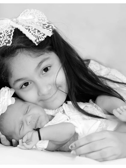A beautiful black and white portrait of an older sister and her newborn sibling. The large bow in the sister's hair adds a charming detail to the photograph.