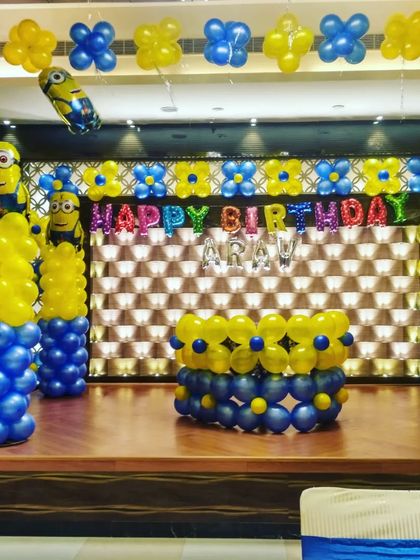 The stage setup for a Minions theme birthday. It includes a custom backdrop, balloon pillars, and a balloon-decorated cake table.