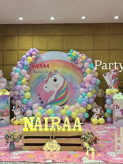 A magical unicorn-themed birthday stage, complete with a rainbow backdrop, pastel balloon arrangements, and multiple unicorn figures. The light-up letters of the name add a sparkling centerpiece.
