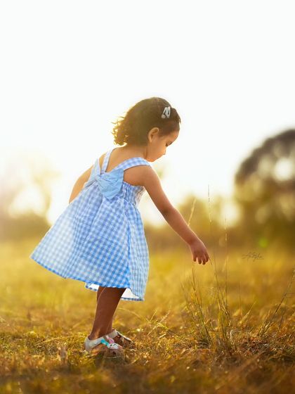 A candid photo of a little girl in a blue gingham dress exploring a field, backlit by the warm afternoon sun.