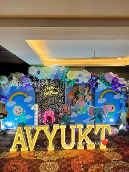 For this grand first birthday, I created a full Cocomelon wonderland. The setup includes a massive backdrop with characters, a silver sequin panel, a balloon arch, large light-up marquee letters spelling the name 'AVYUKT', and multiple character cutouts.