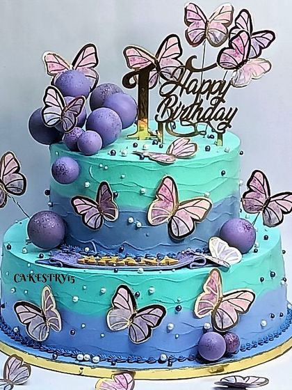 A stunning two-tier butterfly cake in shades of aqua and lavender. The cake is covered in beautiful paper butterflies and decorative purple balls for a vibrant and magical look.