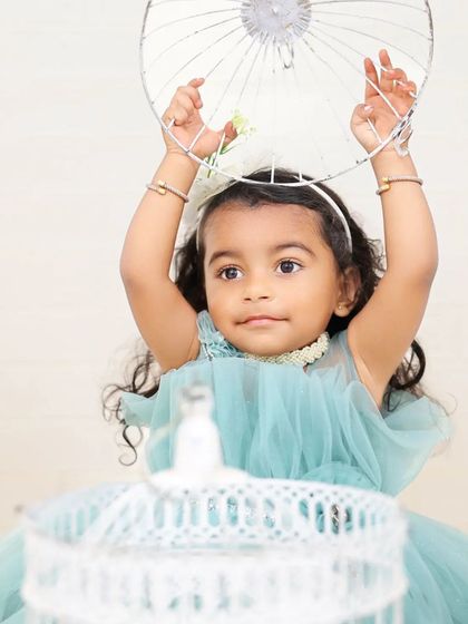 A young girl in a light blue dress plays with a birdcage prop during an outdoor session. The natural background and soft lighting create a dreamy and ethereal portrait.