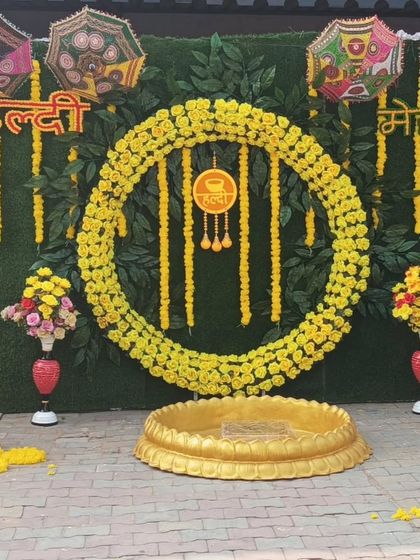 A beautiful Haldi and Mehandi backdrop with a green artificial grass wall. The circular marigold wreath and hanging garlands create a fresh and natural look for your pre-wedding functions.