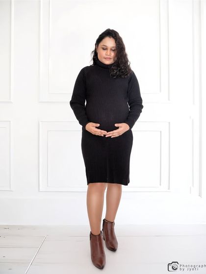 A casual and stylish full-length portrait. The mom-to-be wears a comfortable knit dress and boots for a modern, everyday look.