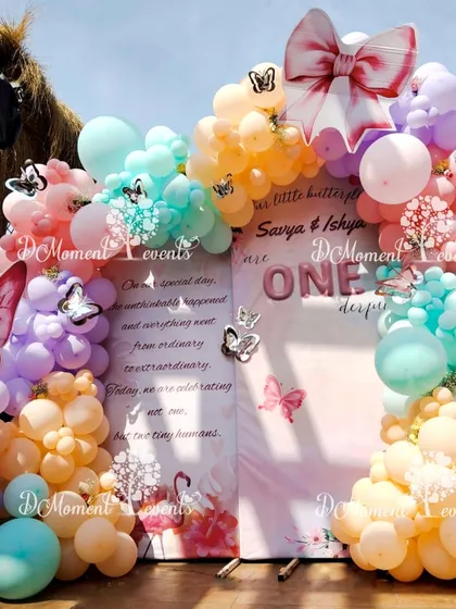 A beautiful photo backdrop for a twins' first birthday. The sign is framed by a cascade of pastel orange, pink, and teal balloons with delicate silver butterfly accents.