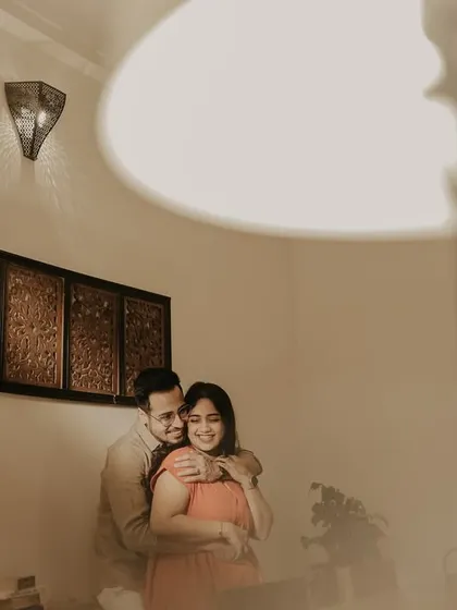 A warm and joyful indoor portrait with the groom embracing the bride from behind. The soft lighting from the lamps creates a cozy and intimate atmosphere for this candid moment.