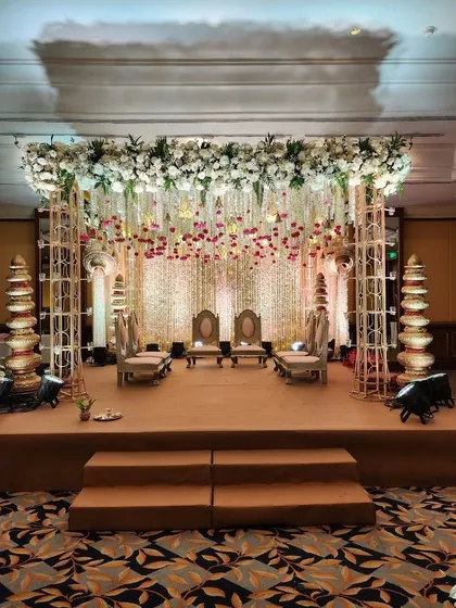 An elegant indoor mandap setup with a metal frame, adorned with a dense canopy of white flowers and hanging floral strings. The traditional stacked 'kalash' elements add a cultural touch.
