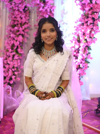 A smiling Buddhist bride against a backdrop of pink flowers. Her hair is styled with soft curls, adding a touch of modern romance to her traditional white saree.