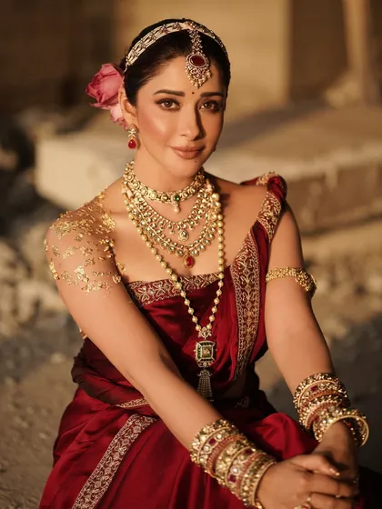 A close-up portrait of Nyra Banerjee, highlighting the intricate gold body art on her shoulder and the detailed temple jewelry. The soft rose in her hair adds a touch of delicate contrast to the powerful look.