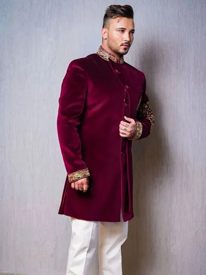 A classic portrait of a male model in a velvet, deep red sherwani. The clean background and studio lighting focus all attention on the rich texture of the fabric and the intricate embroidery on the collar and cuffs.