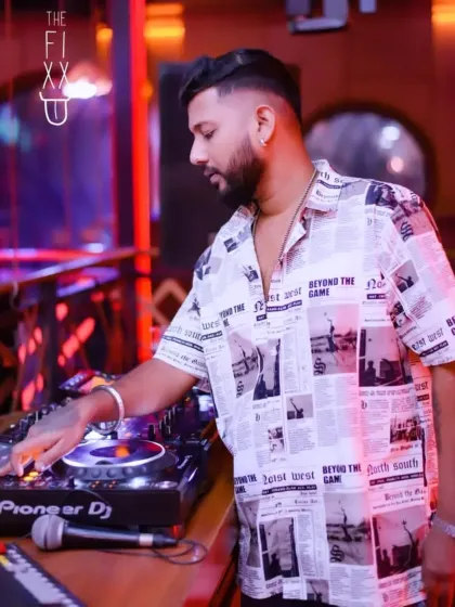 Focused on the Pioneer DJ decks, wearing a unique newspaper-print shirt. Good vibes only.