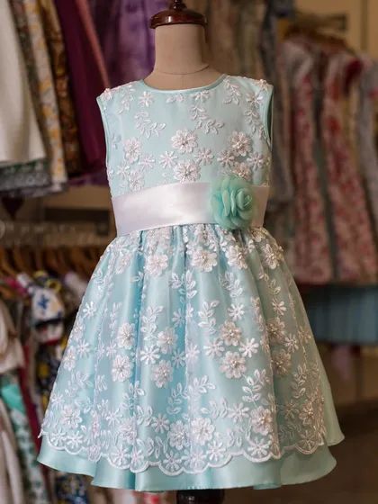 This light blue dress is covered in beautiful white floral lace with pearl centers. A satin sash and a handmade flower complete this incredibly detailed and elegant design.