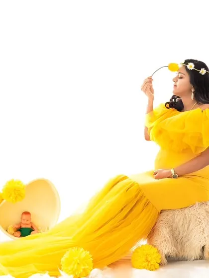 A playful and sweet pose from the sunflower-themed shoot. The expecting mother, seated in her yellow gown, gently smells a yellow flower, adding a touch of whimsy to the bright scene.