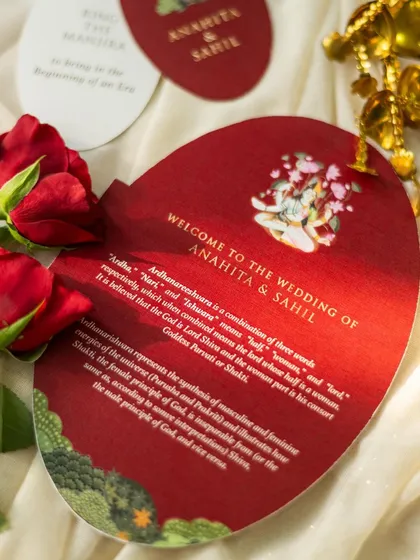 A close-up shot of a red, die-cut wedding invitation, styled with red roses. The dramatic lighting highlights the text and the unique shape of the card.