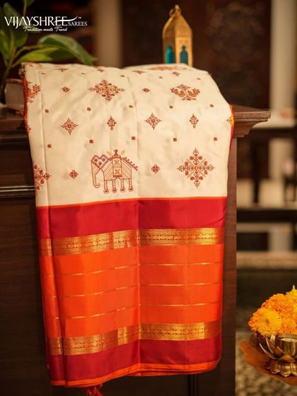 This stunning white Kanjivaram features intricate Kasuthi embroidery from Karnataka, with elephant and geometric motifs, on a bold red and orange gap border.