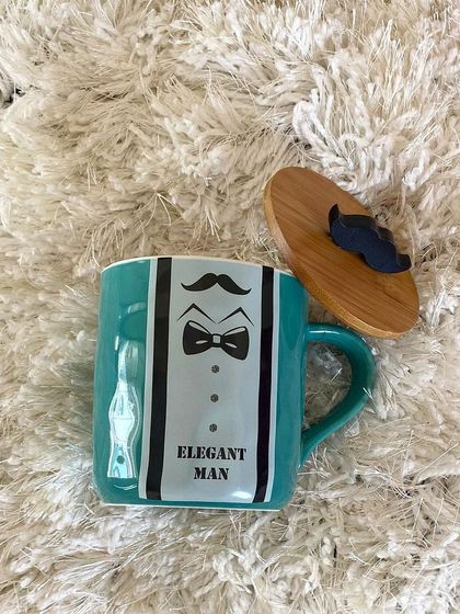 A close up of the teal "Elegant Man" mug. It features a tuxedo graphic and comes with a wooden lid topped with a mustache, combining style with a sense of humor.