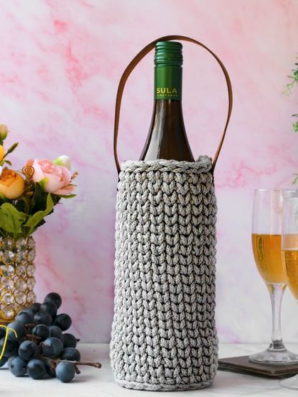 A gray hand-crocheted wine carrier is styled for a celebration, with a bottle of Sula wine, champagne glasses, and grapes against a soft pink marble background.