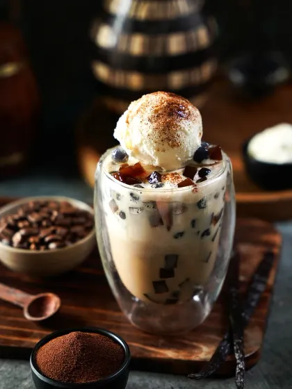 An indulgent coffee-based boba tea topped with a scoop of vanilla ice cream and a dusting of cocoa. The surrounding coffee beans and powder highlight the core ingredients in this moody, atmospheric shot.