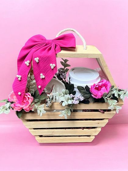 A rustic yet chic wooden crate hamper for her. This gift is adorned with pink flowers and a large pink bow, containing a tumbler and other thoughtful items.
