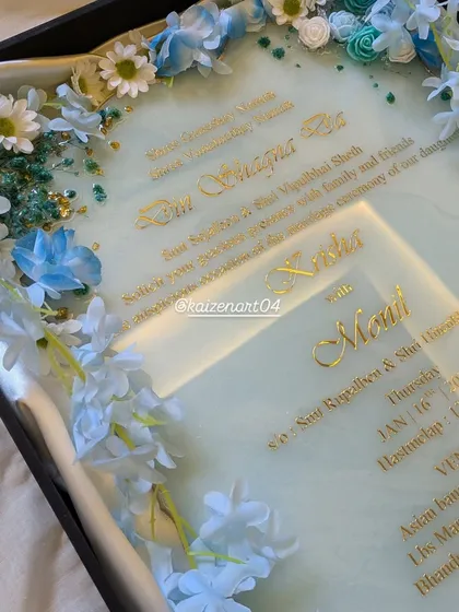 A close-up of the blue resin Kankotri, showing the detail of the metal lettering and the delicate artificial flowers that frame the invitation.