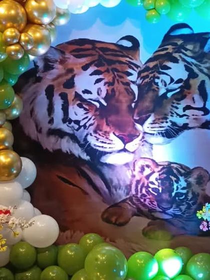 A unique tiger-themed baby shower decoration. The backdrop features a powerful image of a tiger family, surrounded by a green and gold balloon arch and cute teddy bears.