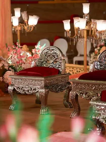 A close-up of the royal seating for the bride and groom within the mandap. The intricately carved silver chairs with red velvet cushions add to the opulent and traditional feel.