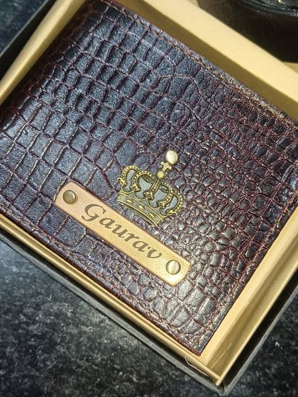 A close-up of a personalized crocodile leather wallet in a unique brown-red shade, featuring a crown charm.
