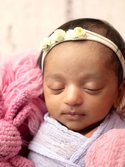 A tiny newborn baby wrapped in soft pink and purple fabrics, nestled in a fluffy basket with a little bunny friend.
