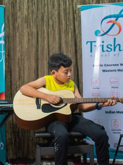 A young student in a yellow shirt plays an acoustic guitar during an open mic session.