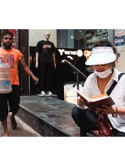 A scene of contrasting activities on a Delhi street. A woman sits absorbed in her book while a laborer walks past carrying a heavy water canister, highlighting the diverse rhythms of city life.