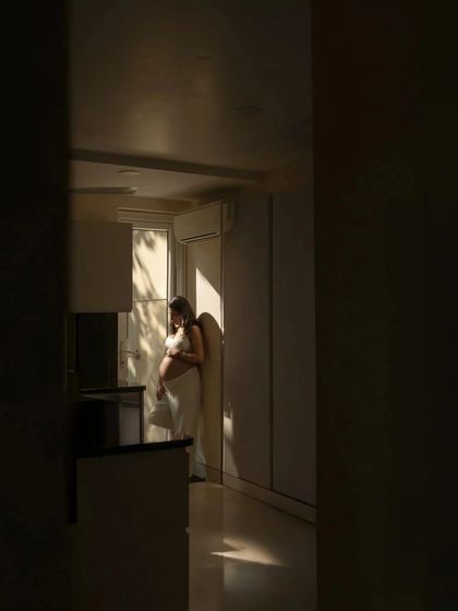 A beautifully composed shot using natural light and shadows. The mother-to-be stands in a sliver of sunlight in her home, creating a dramatic and artistic portrait that feels deeply personal.