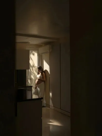 A beautifully composed shot using natural light and shadows. The mother-to-be stands in a sliver of sunlight in her home, creating a dramatic and artistic portrait that feels deeply personal.