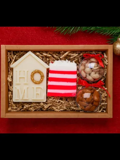 A cozy Christmas-themed gift set. This hamper includes a "Home" soy wax candle, a festive striped soap, and dry fruits, all nestled in a wooden tray.