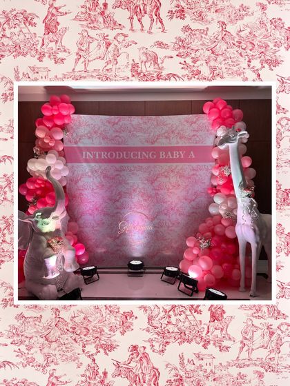 A photo booth backdrop with the text 'Introducing Baby A'. The pink patterned wall is flanked by balloon columns and large animal props for a chic and playful look.
