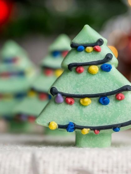 A close-up of a Christmas tree-shaped soap. The selective focus and blurred holiday lights in the background create a magical, festive atmosphere.
