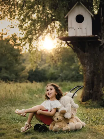 A girl in a red wagon full of teddy bears, set against a magical treehouse backdrop at sunset. A truly whimsical outdoor kid's portrait.