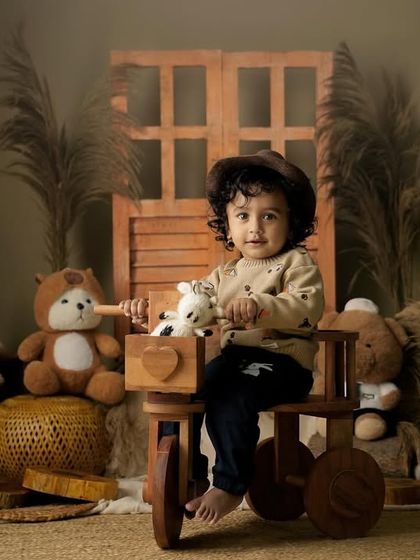 A toddler in a cute hat sits on a wooden tricycle, surrounded by teddy bears in a warm, rustic studio setting.