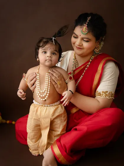 A close-up showing the sweet details of the baby's Krishna costume, from the pearl necklaces to the golden dhoti, as his mother looks on lovingly.