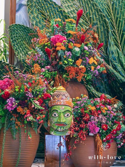 A vibrant floral arrangement featuring a traditional green mask, part of the decor for a Pellikuthuru ceremony.