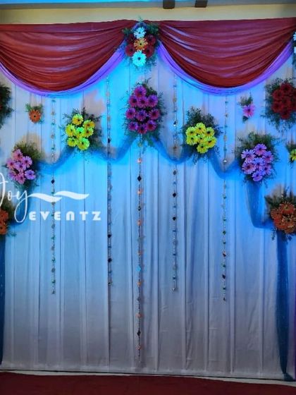 This simple half saree function decoration uses a combination of red and white drapes with colorful flower bouquets. The blue net fabric and hanging crystals add a touch of sparkle to the setup.