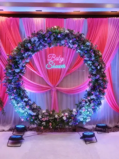 A stunning circular floral arch set against a backdrop of pink and white drapery. The 'Baby Shower' neon sign in the center adds a modern touch to this grand and beautiful stage decoration.