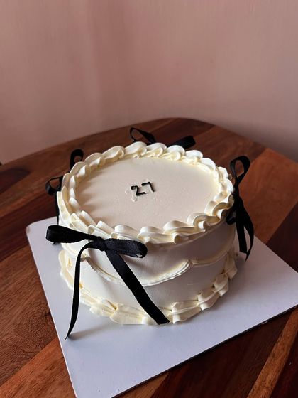 A close-up of a vanilla sponge cake with white chocolate ganache and raspberry jam. The design is Pinterest-inspired, featuring coquette-style piping and black bows.