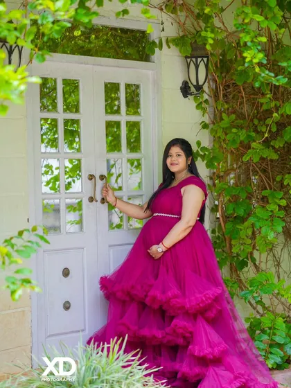 A serene solo portrait of the mother-to-be in a pink gown, standing by a white door surrounded by greenery.