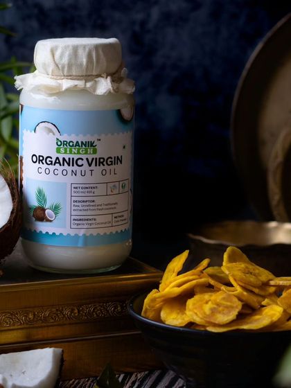 A moody, atmospheric shot of organic virgin coconut oil, styled with crispy banana chips. The dark background and traditional brass elements create a rich, South Indian-inspired aesthetic.